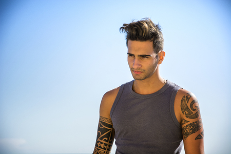 Half Body Shot of a Handsome Athletic Young Man in Trendy Attire, on a Beach in a Sunny Summer Day, Looking Away against Blue Sky Background.の写真素材