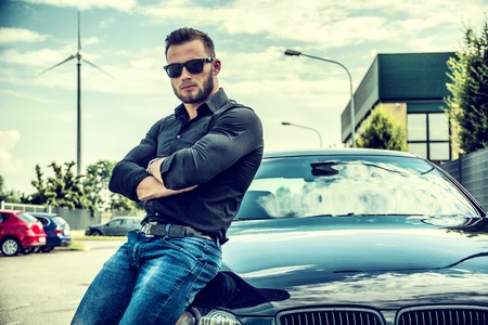 Portrait of brutal young bearded man in sunglasses with crossed arms leaning on his new polished car...