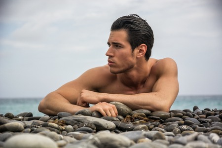 Attractive muscular young shirtless athletic man laying down on pebbles in front of water by sea or ocean shore, looking to a side in a cloudy summer dayの写真素材