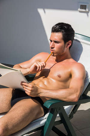 Topless handsome man in panties on deckchair looking pensively away with pencil and notebookの写真素材