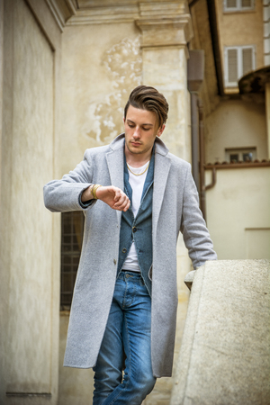 Elegant attractive young man outdoor wearing wool coat, in European city, looking at wrist watchの写真素材