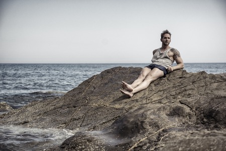 Handsome muscular man on the beach lying on rocks, looking at cameraの写真素材