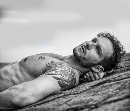 Handsome muscular shirtless man on the beach lying on rocks, looking up to the skyの写真素材