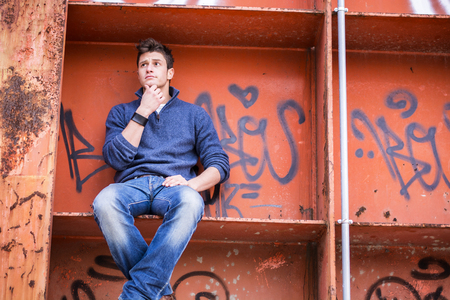 Attractive young man in urban setting sitting on orange metal structure, looking puzzled, thinkingの写真素材