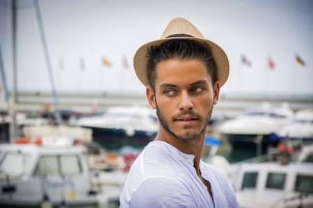 Portrait of handsome young man wearing light brown hat looking calmly away against of harbour with boatsの写真素材