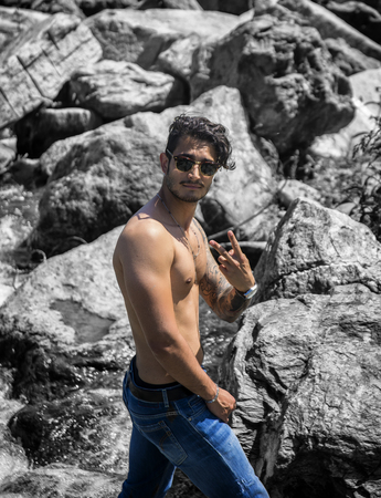 Athletic shirtless young man outdoor at river or water stream, looking away, with rocks and stones in background, doing Victory sign with fingersの写真素材