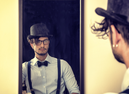 Portrait of brunette young man in glasses, hat, bow-tie, suspenders and shirt looking at himself in a mirror. Vintage lookの写真素材