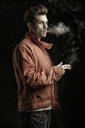 Handsome young man smoking cigarette, dressed as James Dean with red jacket on dark backgroundの写真素材
