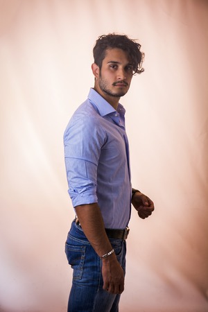 Portrait of brunette young man in light blue shirt and jeans, standing in studio shot against light backgroundの写真素材