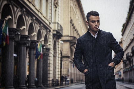 Full length of young man in coat walking under rain along the city street holding cigarette.
