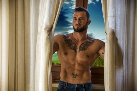 Portrait of sexy shirtless muscular man next to window curtains during the day, wearing only jeansの写真素材