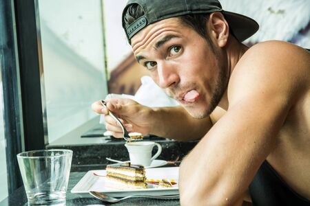 Side view of young trendy man eating dessert in cafe and showing tongue out at camera.の写真素材