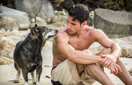 Young topless male in shorts sitting at beach and looking at dog near.の写真素材