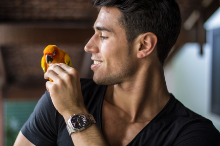 Smiling handsome man feeding parrot sitting on his shoulder. Horizontal indoors shotの写真素材