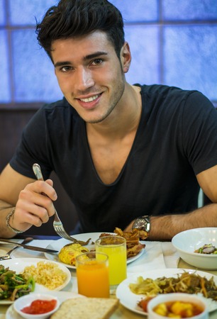 Attractive Young Man Eating Breakfast, Having a Slice of Bread with Jam and Butter and Drinking Fruit Juice, Looking at Camera with a Smileの写真素材