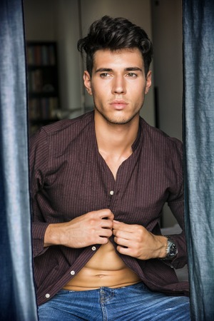 Portrait of sexy young man, buttoning shirt, looking out of window during the dayの写真素材