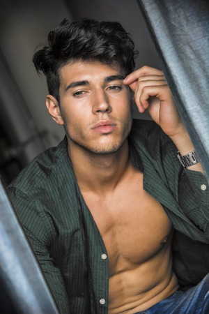 Portrait of sexy young man with shirt open on naked muscular torso, looking out of window during the dayの写真素材