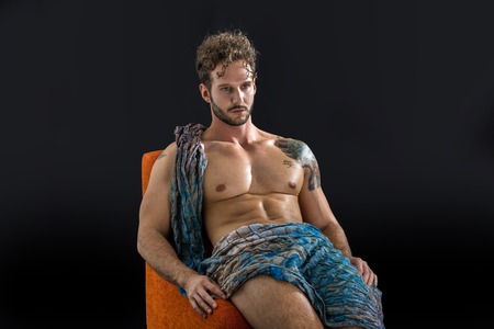 Handsome shirtless muscular man in briefs, covering with long elegant scarf, sitting, isolated on black background in studio shotの写真素材