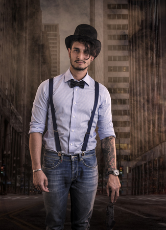 Portrait of brunette young man in glasses, hat, bow-tie, suspenders and shirt on dark smoky city street. Vintage lookの写真素材