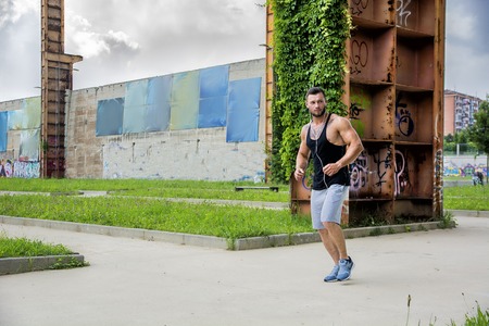 Handsome muscular athletic man running and jogging in city park during the day, wearing black tank...