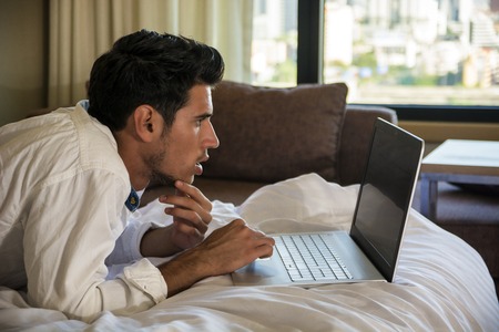Attractive Young Man with Serious Expression, with Laptop on Bed Working on his Start-up Business - Young Male College or University Student Doing Homework, in Bedroomの写真素材