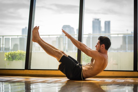 Attractive muscular young man shirtless in gym working out, doing exercises for abs in front of wide windows overlooking modern city skyscrapersの写真素材