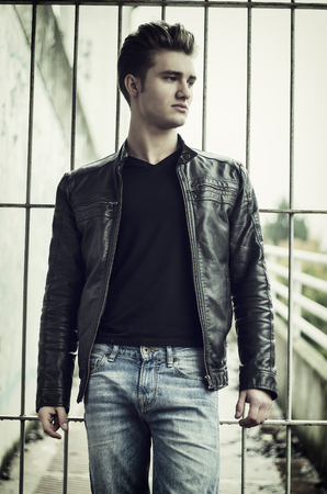 Handsome blond young man outdoors, looking to a side, standing against gateの写真素材