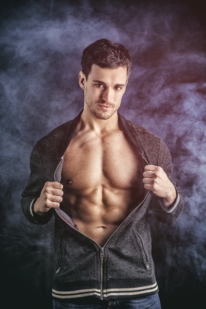 Confident, attractive young man opening vest on muscular torso, ripped abs and pecs. On dark smoky backgroundの写真素材