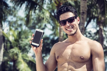 Shirtless Young Handsome Man Showing Cell Phone While Smiling to Camera, with Palm Trees Behind Him....