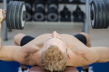Muscular young man shirtless, lifting dumbbells training pecs on gym bench