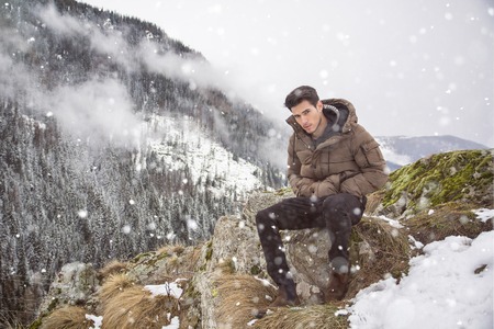 Handsome young man in winter outerwear sitting under snow up the mountain, while looking at cameraの写真素材