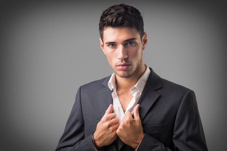 Young businessman confidently posing in front of camera, wearing business suit without neck-tie, with shirt open on neck, on dark backgroundの写真素材