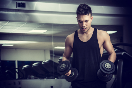 Handsome muscular young man exercising biceps in gym with dumbbellsの写真素材