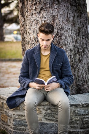 Male student reading book outdoor. Handsome young man reads a book sitting outside on a bench in city parkの写真素材