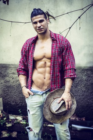 Handsome, muscular young man taking off shirt, wearing jeans and straw hat, outdoorsの写真素材