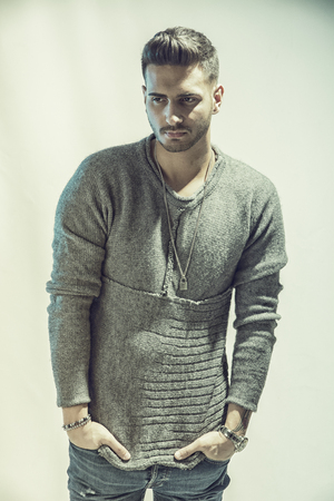Handsome young man wearing grey sweater and ripped jeans, on light background in studio shotの写真素材