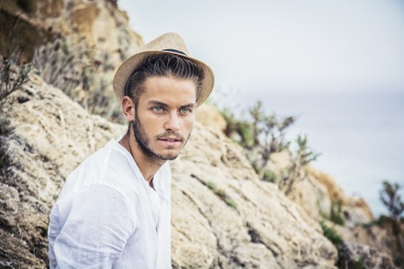 Handsome Young Man in Trendy Attire, on a Beach in a Sunny Summer Day, Wearing a White Shirt and Straw Hatの写真素材