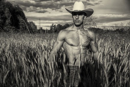 Portrait of sexy farmer or cowboy in hat looking to a side, while standing next to hay field in countrysideの写真素材