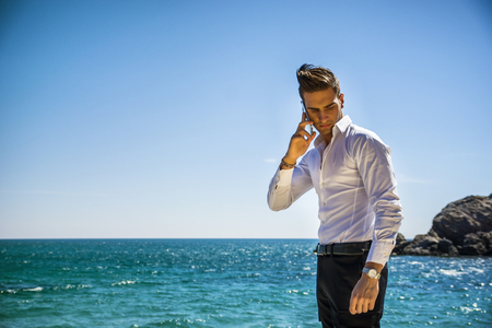 Young man at the seaside talking on cell phone while looking at the sea. Large copyspace. Sea waves on backgroundの写真素材