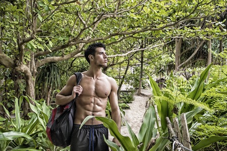 Young muscular topless man with backpack and phone looking away on background of tropical forest.の写真素材
