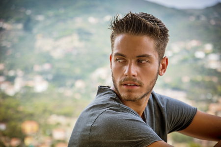Handsome Young Man in Trendy Attire, in a Sunny Summer Day with Italian Sea Coast in the Distance, Wearing a White Shirtの写真素材