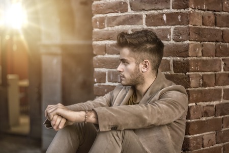 Attractive young man sitting against brick wall, looking away to a sideの写真素材