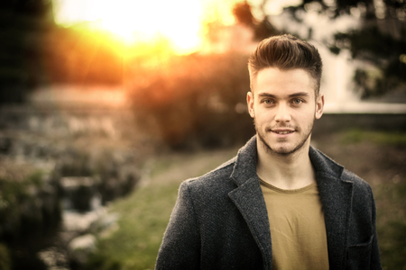 Handsome young man standing in city park, looking confident and relaxed in autumn or spring evening