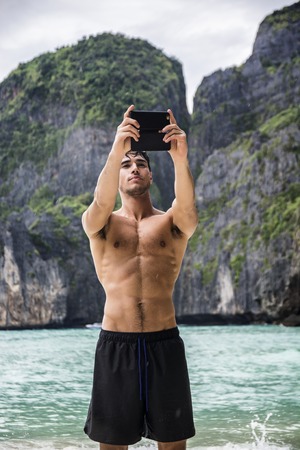 Half body shot of a handsome young man using cell phone to take photo, standing on a beach in Phuket Island, Thailand, shirtless wearing boxer shorts, showing muscular fit bodyの写真素材