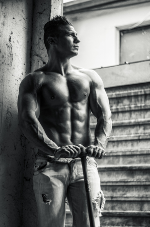 Muscular shirtless young man holding farming tool in his hands, sweaty in jeansの写真素材