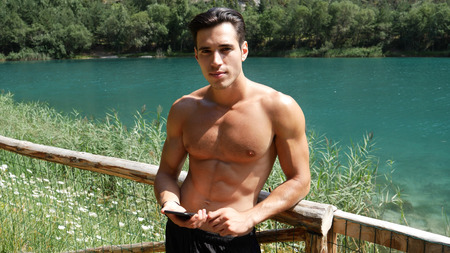 Young shirtless muscular handsome man with smartphone typing on it or using it to go online, at lake in forestの写真素材