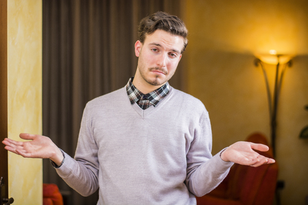 Confused or doubtful young man shrugging with palms open, indoor shot in a houseの写真素材