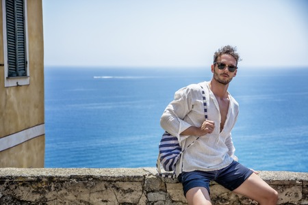 Handsome fit man sitting on old brick wall in Italian town, with the sea far behind him. Horizontal outdoors shotの写真素材