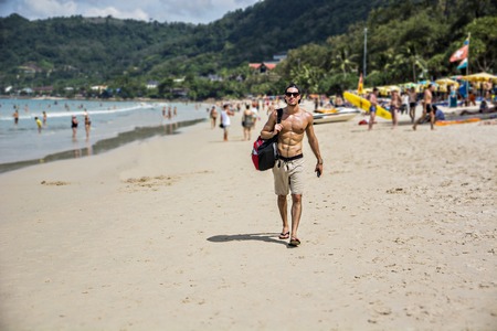 Full body shot of a handsome young man walking on a beach in Phuket Island, Thailand, shirtless wearing boxer shorts, showing muscular fit body. Full length shot.の写真素材