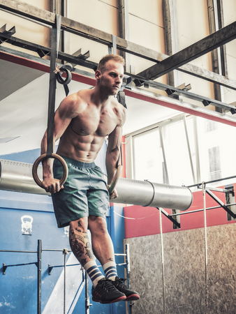 Handsome muscular young man athlete exercising in gym, hanging and pushing up with rings. Tattoo...